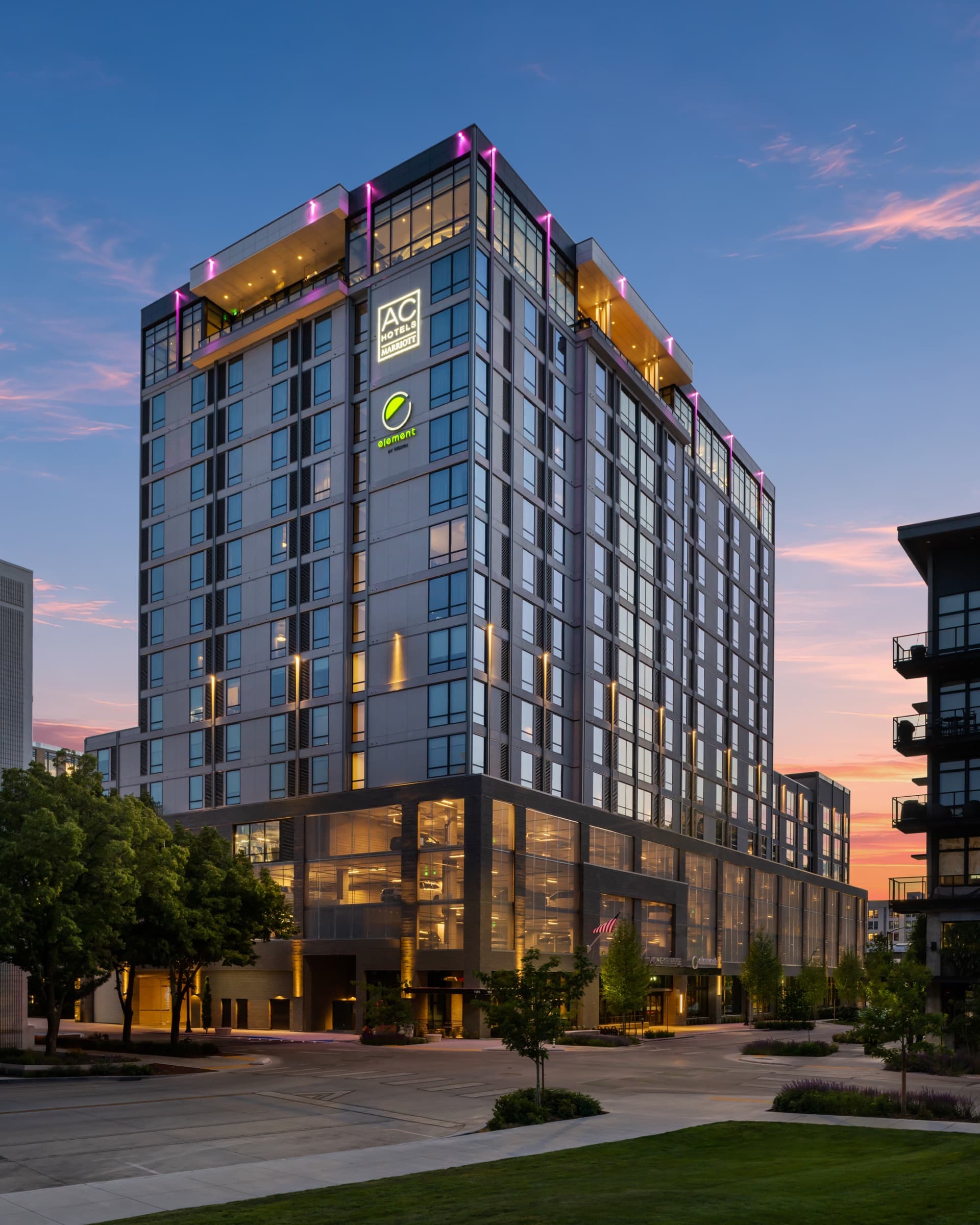 AC Hotel and Element Boise at dusk, seen from street level — both signs lit, the podium glass glowing
